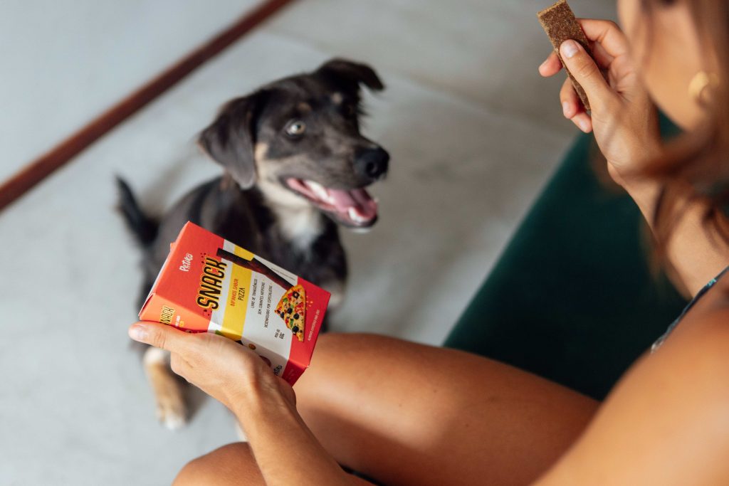 Tutora segurando Snack – Petisco sabor pizza e seu cachorro sentado olhando para esse petisco