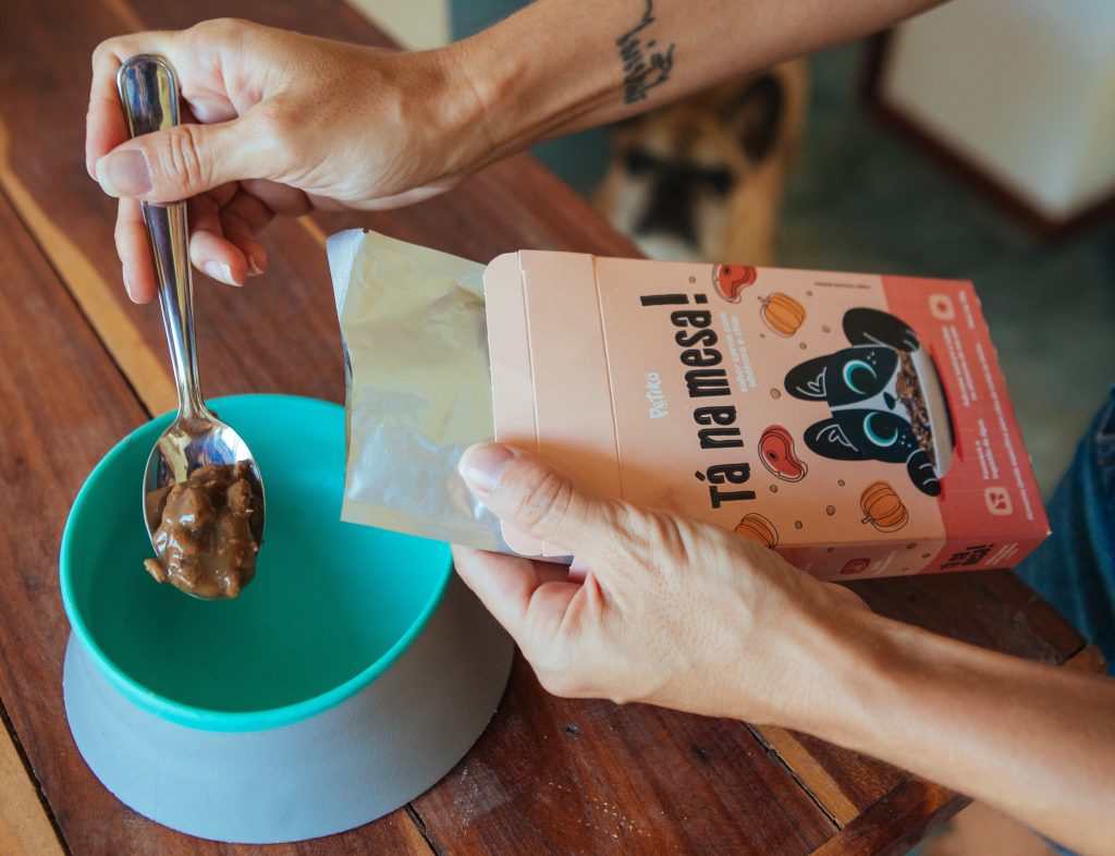 Tutora servindo o alimento úmido Tá na Mesa, enviado no BOX.Petiko, em um comedouro azul para seu cachorro que a observa